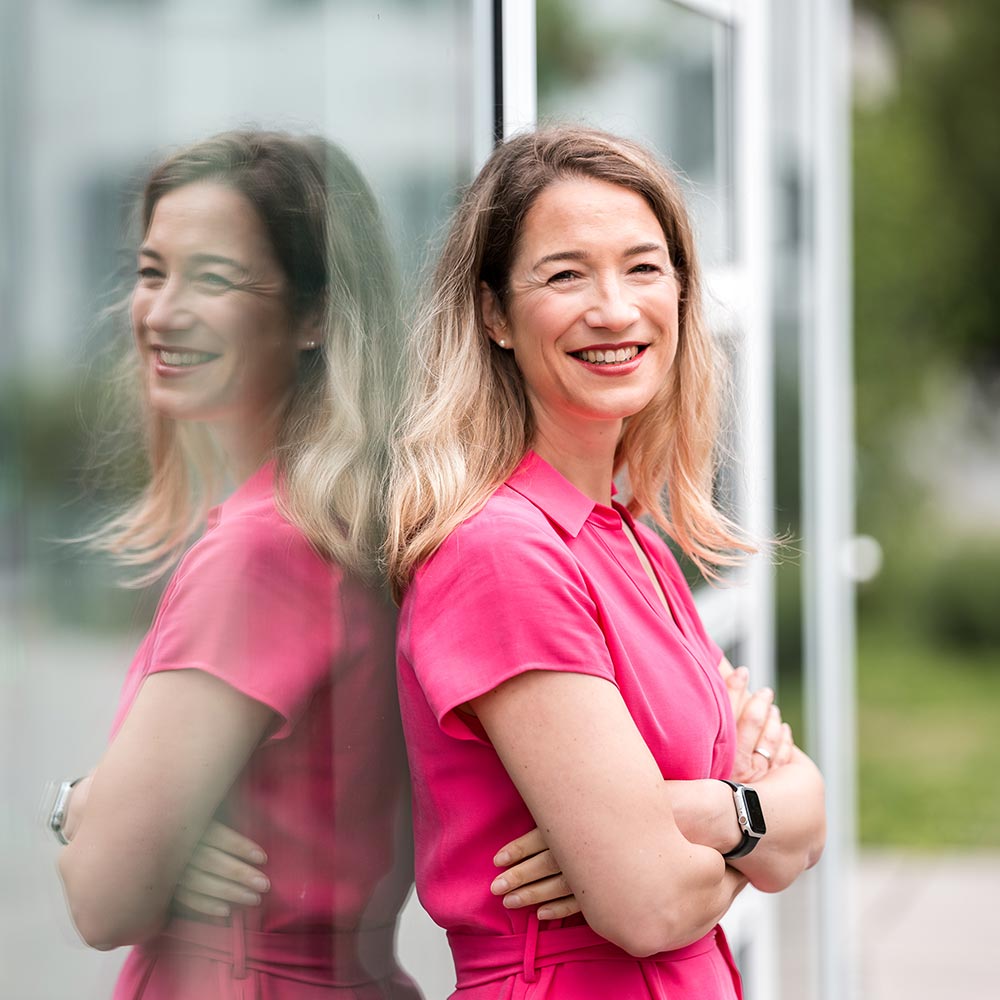 Anne Hegemann, Geschäftsführung FREIBADEN: Junge, blonde Frau steht mit verschränkten Armen angelehnt an eine Glasscheibe. Sie trägt einen pinken Jumpsuit und lächelt freundlich in die Kamera.